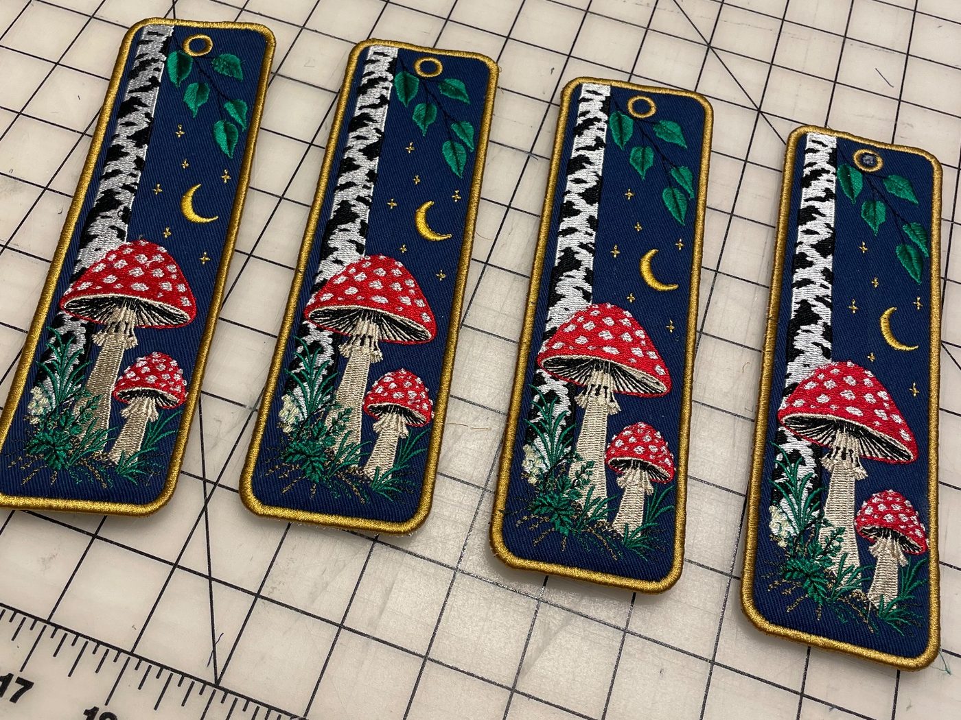 Four finished Beneath the Birch bookmarks side by side on a cutting mat: each has a white birch trunk, green leaves, a gold crescent moon and stars on a navy ground, two red fly agarics with white spots among ferns, and a gold border.