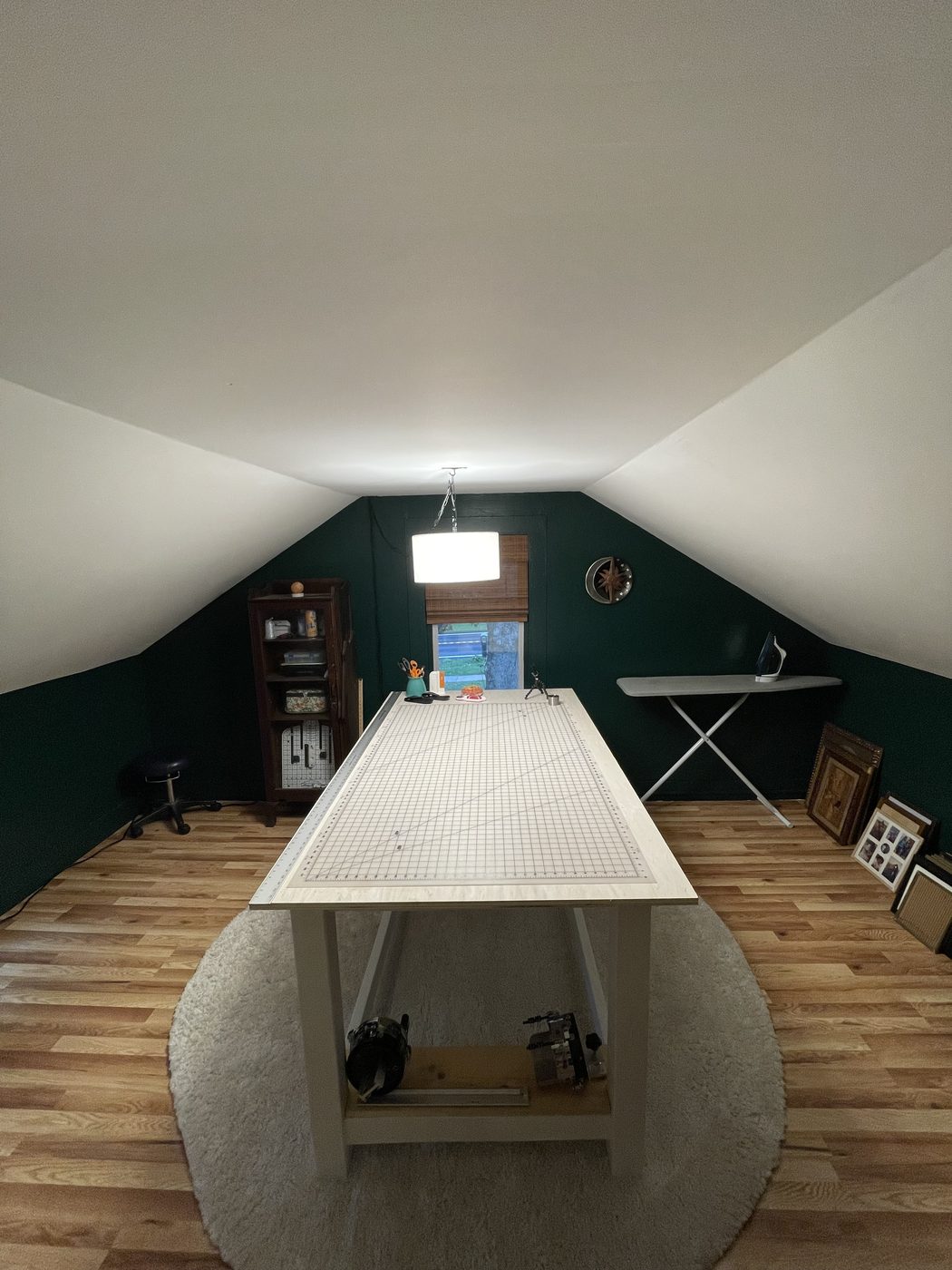 The same sloped-ceiling room as the studio: deep green walls, wood floors, a long white cutting table with a gridded mat on a round white rug under a drum pendant light, a glass-front cabinet of supplies, an ironing board, and framed pictures leaning against the wall.