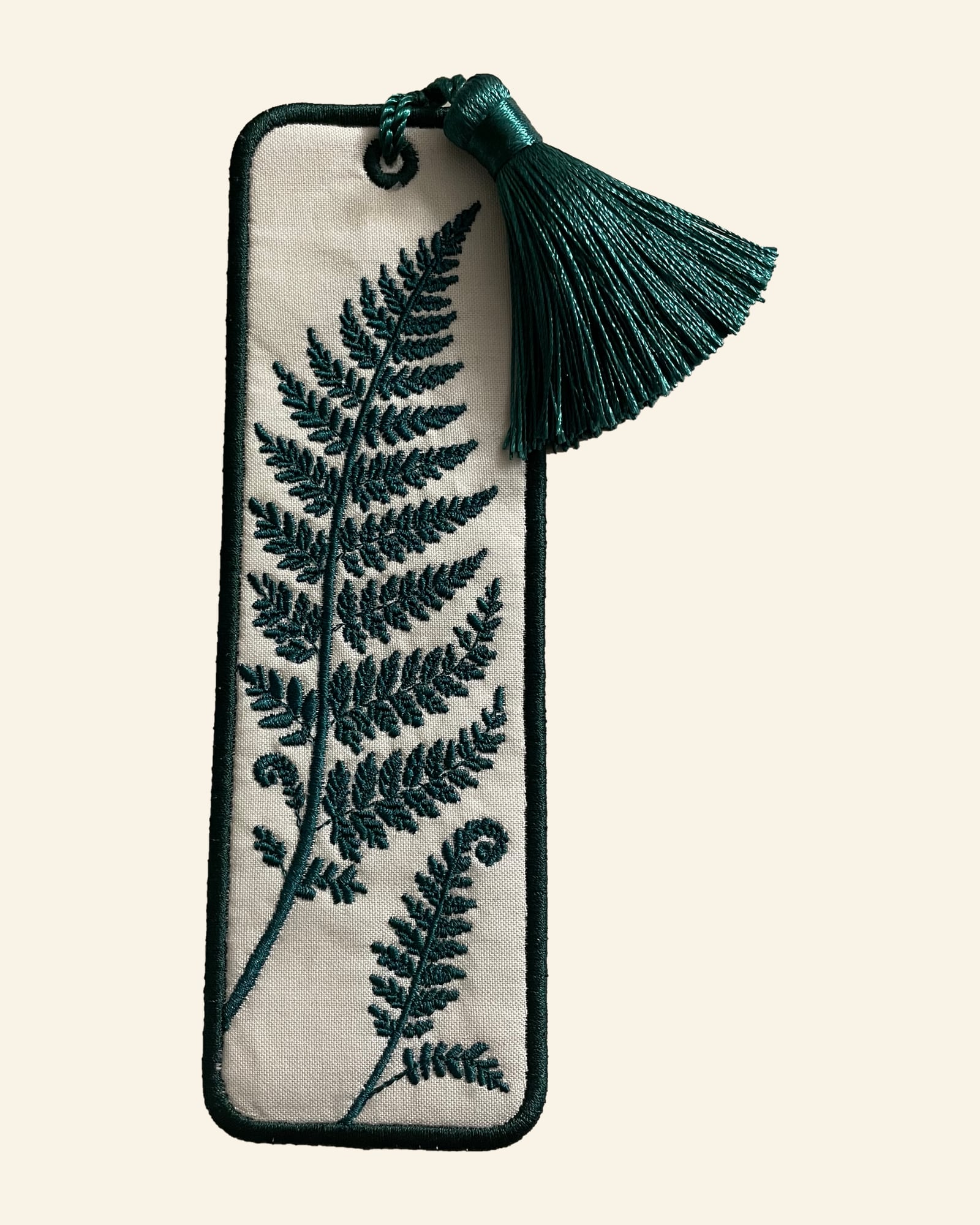 The Fern & Frond bookmark: a cream bookmark with a deep green border, embroidered with a tall fern frond and a smaller frond with curled fiddleheads, a green tassel at the top.